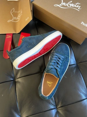 Blå mockasneakers från Louboutin - Snygga blå sneakers i mocka från Christian Louboutin med klassisk röd sula och vita kanter. Skorna har lågt skaft, matchande blå skosnören och diskreta sömdetaljer. Perfekta för dig som vill sticka ut med exklusiv stil. HELT NYA ALDRIG ANVÄNDA 10/10 skick. Alla tillbehör medföljer. Storlek 43 och 45 finns - OBS TAR SLUT SNABBT