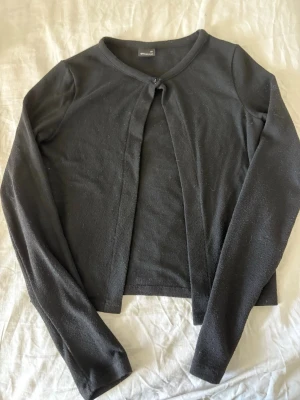Svart kofta från Gina Tricot - Säljer en svart, tunn kofta från Gina Tricot i storlek XS. Koftan har lång ärm, rund halsringning och stängs med en knapp upptill. Perfekt att slänga över en topp för en enkel och stilren look.