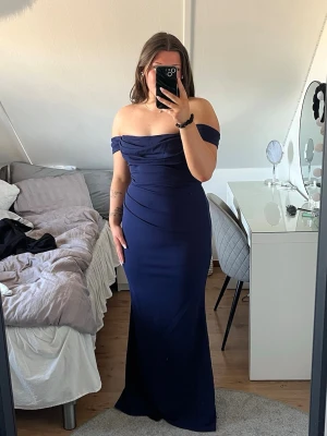 Marinblå offshoulder långklänning - Elegant marinblå långklänning med offshoulder-design och figurnära passform. Klänningen har draperade detaljer över bysten och höfterna som ger en snygg siluett. Perfekt för speciella tillfällen som bal. Köpt från Bubbleroom och är ej använd så lappen hänger kvar 