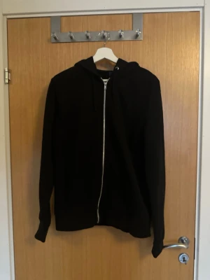 Svart zip-hoodie med huva från divided - En klassisk svart hoodie med dragkedja framtill och huva från divided. Hoodien har långa ärmar och ribbade muddar vid ärmslut och nederkant. Perfekt för en chill och avslappnad stil. Storlek M. Materialet känns mjukt och bekvämt mot huden.