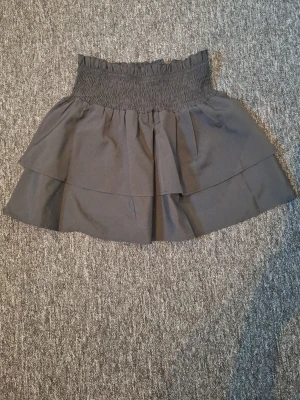 Svart volangkjol med smock - Snygg svart kortkjol med bred smock i midjan och dubbla volanger. Kjolen har en feminin och trendig look, perfekt att matcha med en enkel topp.