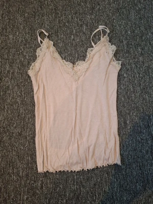 Beige ribbad linnetopp med spets - Söt beige linnetopp med tunna axelband och v-ringning dekorerad med spets. Toppen är ribbad och har en feminin, mjuk look som passar perfekt till jeans eller kjol. Lätt och luftig för varmare dagar. Ps lappen är avklippt 