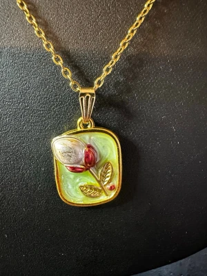 Guldhalsband med blomma och blad - Unikt halsband med guldfärgad kedja och rektangulärt hänge. Hänget har en relief av en blomknopp i silver och rött, gröna blad och en ljusgrön bakgrund. Perfekt för dig som gillar detaljerade och färgglada smycken.
