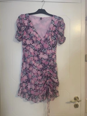 Blommig kortklänning från Divided - Superfin kortklänning från Divided med blommigt mönster i rosa, lila och vitt. Klänningen har v-ringning, korta puffärmar och snörning nertill som ger en snygg rynkad effekt. Perfekt för sommarens alla tillfällen!