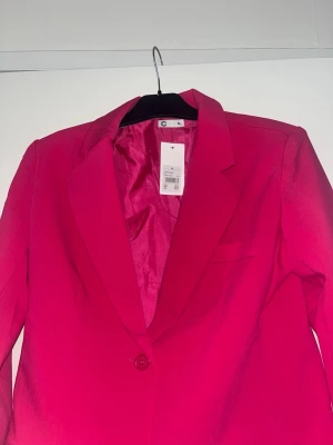 Ceriserosa kavaj från Cubus - Stilren ceriserosa kavaj från Cubus med klassisk krage, en knapp framtill och två fickor. Kavajen har en figurnära passform och är helfodrad för extra komfort. Perfekt för att lyfta vilken outfit som helst med en färgklick.