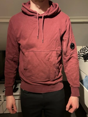 Vinröd hoodie från C.P. Company - Säljer en vinröd hoodie från C.P. Company med klassisk huva och dragsko, samt en ficka framtill. Ärmen har den ikoniska linsen på sidan. Tillverkad i mjuk bomull, perfekt för en avslappnad och snygg stil.