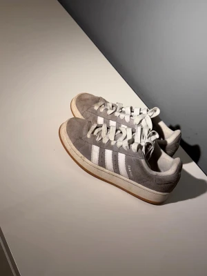 Grå Adidas Campus sneakers - Säljer ett par grå Adidas Campus sneakers med klassiska vita ränder på sidorna och vit sula. Ovandelen är i mocka och skorna har vita skosnören. Perfekta för dig som gillar en clean och tidlös stil.