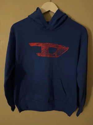 Diesel hoodie mörkblå med röd logo Y2K - Snygg blå hoodie från Diesel med stor röd logga på bröstet. Tillverkad i 100% bomull för skön känsla och har klassisk känguruficka framtill. Perfekt för dig som vill ha en streetwear-look med edge.