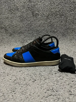 Saint Laurent court classic SL/10 - Snygga sneakers från Saint Laurent i svart och blått läder med vita sulor. Klassisk låg modell med svarta skosnören och diskret logga på hälen. Perfekta för dig som gillar stilrena men ändå färgstarka skor.