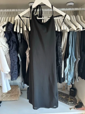 Svart omlottklänning med tunna band - Säljer en svart kortklänning med tunna axelband och omlottdetalj i ryggen. Klänningen har en enkel, rak passform och är tillverkad i ett lätt, slätt material som ger en stilren look. Perfekt för dig som gillar minimalistisk stil.