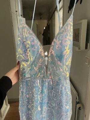 Ljusblå balklänning/festklänning  - Magisk ljusblå långklänning med glittrigt mönster och djup v-ringning. Klänningen har tunna axelband, slits framtill och skimrande detaljer.  Perfekt för bal eller festliga tillfällen. Den är dessutom helt ny och oanvänd.