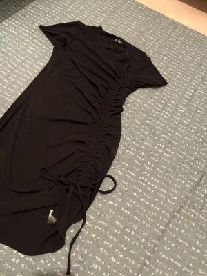 Svart kort klänning från H&M  - Svart kort klänning från H&M med snörning längs sidan som ger en snygg rynkad effekt. Klänningen har korta ärmar och är gjord i mjukt, stretchigt material som sitter bekvämt. Perfekt för dig som gillar enkel men trendig stil.