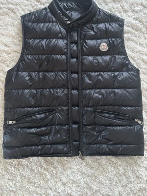 Svart dunväst från Moncler - Snygg svart dunväst från Moncler med glansig finish och klassisk logga på bröstet. Västen har två dragkedjeförsedda fickor framtill och hög krage med tryckknapp. Perfekt för lager-på-lager och riktigt stilren look.  Det finns en liten lagning på ryggen eftersom det var ett lite hål där syns ytterst lite.