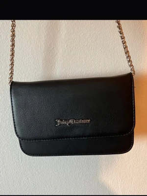 Svart axelväska Juicy Couture - Snygg svart axelväska från Juicy Couture i slätt konstläder med silverfärgad kedjerem och logga framtill. Väskan har en stilren, rektangulär form och passar perfekt för dig som gillar en clean och trendig look.