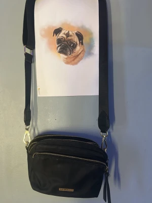 Svart axelväska från Don Donna - Snygg svart axelväska från Don Donna med justerbar axelrem och två stora fack med guldfärgade dragkedjor. Väskan har en stilren design i slätt material och en liten guldfärgad logga framtill. Perfekt för att bära det viktigaste med stil.