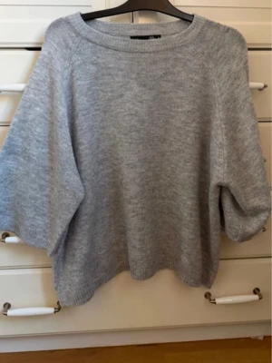 Ljusgrå stickad tröja 157 - Mysig ljusgrå stickad tröja från 157 med rund halsringning och avslappnad passform. Tröjan har breda ärmar och ribbade muddar vid halsen. Perfekt för lager på lager och enkel att styla till olika outfits.