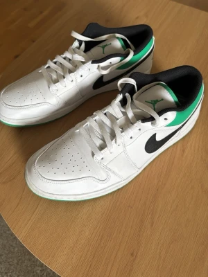 Air Jordan 1 Low vit/grön/svart - Säljer ett par Air Jordan 1 Low sneakers i vitt läder med svarta detaljer och gröna inslag på häl och yttersula. Skorna har blivit använd 1 gång så mindre tecken på användning.