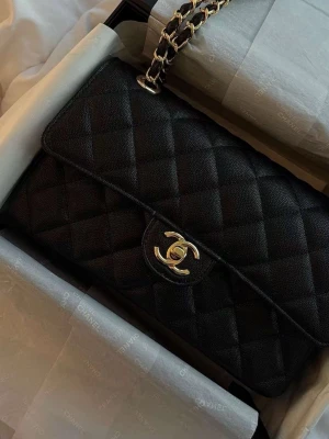 Svart quiltad handväska - Säljer en klassisk svart quiltad handväska med guldfärgad kedjerem  och ikonisk logga framtill. Väskan är tillverkad i läder och har en elegant, tidlös design som passar perfekt till alla tillfällen. Lådan och presentpappret samt dustbag medföljer. 