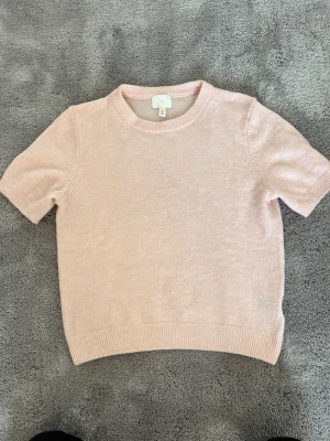 Ljusrosa stickad t-shirt från H&M - Söt ljusrosa stickad t-shirt från H&M med korta ärmar och rund halsringning. Tröjan har ribbade kanter vid hals, ärmslut och nederkant. Perfekt för en avslappnad och trendig look.