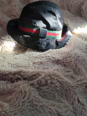 Svart bucket hat - Snygg svart bucket hat med klassiskt grönt och rött band runtom. Hatten är tillverkad i Italien och har en mjuk, lätt glansig yta. Perfekt accessoar för dig som vill sticka ut med en ikonisk och trendig look. Står storlek m men uppskattar en xs-s