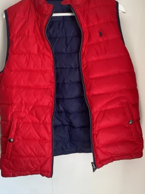 Röd dunväst från Polo Ralph Lauren - Snygg röd dunväst från Polo Ralph Lauren med marinblått foder och diskret logga på bröstet. Västen har två fickor med knappstängning och dragkedja framtill. Perfekt för lager på lager och ger en sportig vibe.