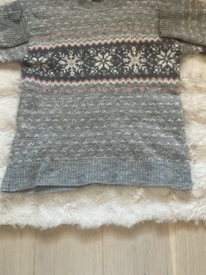 Grå stickad tröja med snöflingor - Mysig grå stickad tröja med vita och rosa snöflingor och zigzag-mönster över bröstet. Tröjan har rund halsringning, långa ärmar och ribbstickade muddar. Perfekt för kalla dagar och har en klassisk vinterlook.