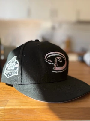 Svart Arizona Diamondbacks keps - Snygg svart keps från New Era med Arizona Diamondbacks-logga broderad framtill i rosa och vitt. På sidan finns ett 20th Anniversary-märke. Klassisk flat brim och justerbar baksida. Perfekt för dig som gillar sportig streetstyle och vill sticka ut.