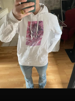 Vit hoodie med rosa tryck - Säljer en vit hoodie med huva och magficka. På bröstet finns ett stort rosa och lila abstrakt tryck som ger en unik vibe. Hoodien har dragsnören i huvan. 