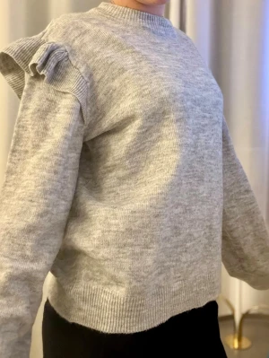 Ljusgrå stickad tröja med volang - Mysig ljusgrå stickad tröja med volangdetalj på axlarna och ribbade muddar vid ärmslut och nederkant. Tröjan har en rund halsringning och är perfekt för lager-på-lager-stil under kyliga dagar.