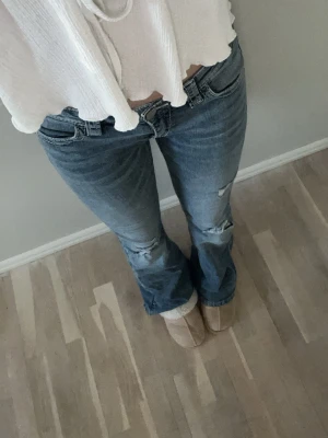 Blå bootcut jeans med slitningar - Säljer ett par blå bootcut jeans med låg midja och slitningar på knäna. Jeansen är tillverkade i denim med lätt stretch. Perfekta för en avslappnad och trendig look. Skriv vid funderingar!❤️