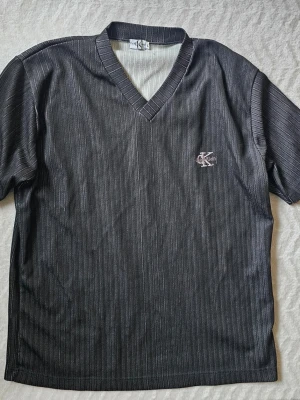 Svart v-ringad topp från Calvin Klein - Snygg svart topp från Calvin Klein Jeans med diskreta vita ränder och v-ringad hals. Kortärmad modell med broderad CK-logga på bröstet. Perfekt för en clean och stilren look.