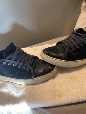 Lanvin marinblåa  sneakers i mocka och skinn - Snygga marinblåa  sneakers från Lanvin med ovandel i mocka och detaljer i blankt skinn på tån. Skorna har mörkblå snörning, metallöljetter och en kontrasterande beige sula. Perfekta för dig som gillar stilrena och exklusiva sneakers. Sulan är mycket finare i verkligheten. Storlek UK 7 vilket motsvarar 42/43