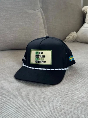 Svart keps Golfio.se Eat Sleep Golf - Svart keps från Golfio.se med patch framtill där det står 'Eat Sleep Golf Repeat'. Kepsen har vit och svart snodd över skärmen, svensk flagga på sidan och ventilerande mesh på bakpanelen. Broderad logga på sidan. Justerbar baktill.