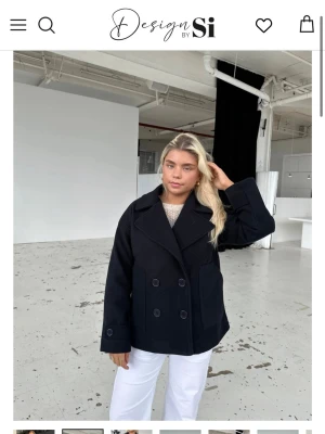 Svart kort kappa från Design by Si - Stilren svart kappa med dubbelknäppning och bred krage från Design by Si. Jackan har två stora framfickor och en rak, något boxig passform. Perfekt för dig som gillar klassisk men trendig stil.