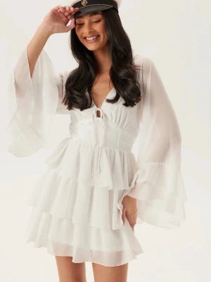 Vit volangklänning - Supersöt vit kortklänning med flera volanglager och vida, transparenta ärmar. Djup v-ringning med knyt framtill och omlottkänsla. Tillverkad i ett lätt och luftigt material. Köpte nyligen men beställde fel storlek, så helt oanvänd med lappar kvar. Jätte fin klänning till tex student eller sommarfest! 