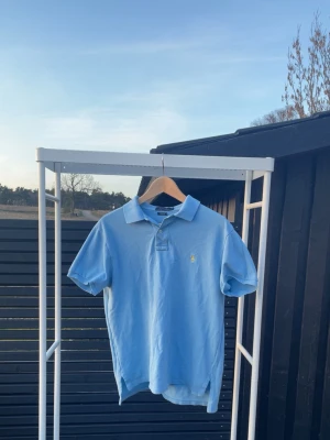 Ljusblå pikétröja från Ralph Lauren - Klassisk ljusblå pikétröja från Ralph Lauren med liten gul broderad logga på bröstet. Tröjan har korta ärmar, krage och knappar vid halsen. Perfekt för en avslappnad och stilren look.