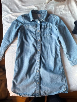 Ljusblå jeansklänning med knappar - Snygg ljusblå jeansklänning från H&M med långa ärmar och knappar hela vägen framtill. Klänningen har två bröstfickor och klassisk skjortkrage. Perfekt för dig som gillar en avslappnad men trendig stil.