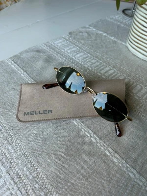 Ovala solglasögon från Meller - Snygga ovala solglasögon från Meller med tunna guldfärgade metallbågar och mörkgröna/svarta glas. Skalmarna har sköldpaddsmönster och glasögonen är oanvända. Fodral och putsduk medföljer. 
