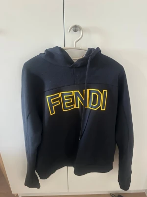 Mörkblå hoodie från Fendi med tryck - Säljer en mörkblå hoodie från Fendi med stor gul FENDI-logga på bröstet. Hoodien har huva med dragsko och långa ärmar. Tillverkad i mjukt material som känns skönt mot huden. Perfekt för dig som vill ha en stilren och trendig look.