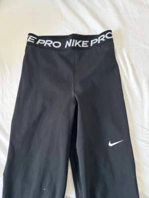 Svarta Nike Pro leggings - Säljer ett par svarta Nike Pro leggings med bred resår i midjan där det står 'NIKE PRO' i vitt. De har en tight passform och är tillverkade i ett stretchigt material som passar perfekt till träning. Klassisk Nike-logga på benet.