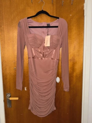 Rosa långärmad omlottklänning - Supersnygg rosa klänning från Oh Polly med lång ärm och omlottdetaljer. Klänningen har markerad byst, mesh-tyg och draperad kjol som ger en riktigt trendig look. Perfekt för dig som vill sticka ut med en feminin och modern stil.