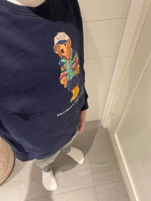 Mörkblå Polo Bear sweatshirt - Mörkblå sweatshirt från Ralph Lauren med ikoniska Polo Bear-trycket framtill. Björnen har färgglada kläder och keps, vilket ger tröjan en lekfull och trendig vibe. Rund halsringning och långa ärmar. Perfekt för dig som gillar streetwear och klassiska märken.