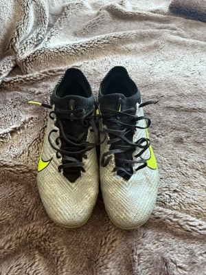 Nike Mercurial silvriga  fotbollsskor - Säljer ett par Nike Mercurial fotbollsskor i grått med svarta detaljer och neongula swoosh-loggor. Skorna har snörning, strumpliknande skaft och är tillverkade i ett lätt syntetmaterial för bra bollkontroll och grepp på planen.