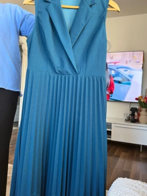 Blå plisserad långklänning från Zara - Snygg blå långklänning från Zara med plisserad kjol och ärmlös design. Klänningen har en markerad krage och v-ringad omlott upptill, vilket ger en elegant look. Perfekt för dig som vill sticka ut med stilrena detaljer.
