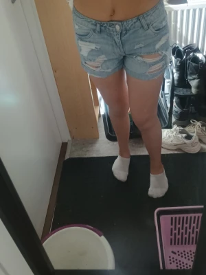 Ljusblå denimshorts med slitningar - Säljer ett par ljusblå denimshorts med slitna detaljer och fransig kant. Shortsen har fem fickor, knappgylf och är högmidjade för en snygg passform. Perfekta till sommaren och ger en avslappnad look.