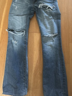 Low waist bootcut jeans - Säljer ett par blå lågmidjade bootcut jeans med slitningar och hål på knäna. Jeansen har ljus tvätt och detaljerade bakfickor med broderi. Perfekta för en avslappnad och trendig look. Skriv privat för mått! Storleken är w26 vilket motsvarar xs/s, pris kan också diskuteras!