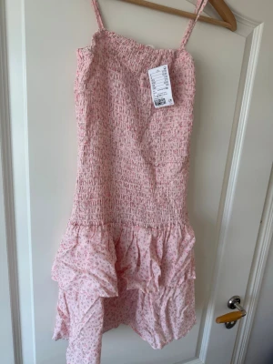 Rosa blommig volangklänning H&M - Söt rosa kortklänning från H&M med smala axelband och volangkant nedtill. Klänningen har ett smockat liv och ett ljust blommigt mönster. Klänningen kan också fungera som en tube klänning. Passar perfekt för de varma dagarna som kommer☀️💕. (Storleken är 12-13 men passar xs också.)