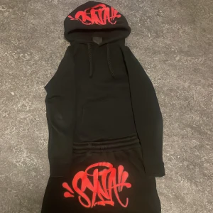 Syna world set - Säljer en svart hoodie med huva och dragsnören. På huvan och byxorna finns stora röda graffiti-tryck med texten 'SNA'. Hoodien har en avslappnad passform och är tillverkad i mjukt material, perfekt för en streetwear-look.