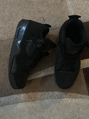 Svarta Air Jordan 4 Retro sneakers - Säljer ett par helsvarta Air Jordan 4 Retro sneakers med klassisk meshpanel på sidorna, plastdetaljer och tjock sula. Skorna har högt skaft och ikonisk design som passar perfekt till streetwear. Materialet är en mix av läder och syntet.