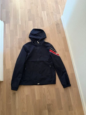 Marinblå Moncler vindjacka med huva - Snygg marinblå vindjacka från Moncler med huva och dragkedja framtill. Jackan har en röd, vit och svart rand på ena ärmen samt Moncler-logga. Tillverkad i lätt och tåligt material, perfekt för blåsiga dagar.